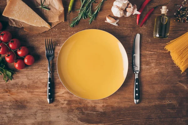 Vista superior de la pasta de fila, ingredientes frescos y plato con cuchillo y tenedor en la mesa de madera - foto de stock