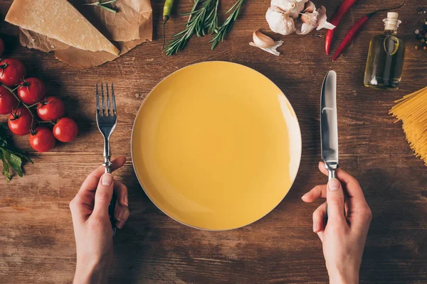 Vista recortada de las manos con cuchillo y tenedor en el plato con pasta de fila e ingredientes frescos alrededor de la mesa de madera - foto de stock