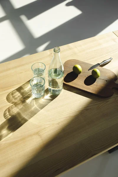 Overhead view of limes on wooden board and water in glasses and bottle on wooden table — Stock Photo