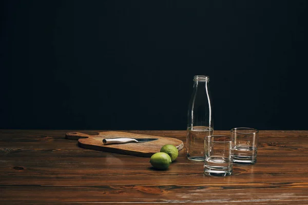 Wooden board with knife, limes and water on brown tabletop — Stock Photo