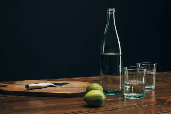 Tabla de madera con cuchillo, limas y botella de agua en la mesa de madera - foto de stock