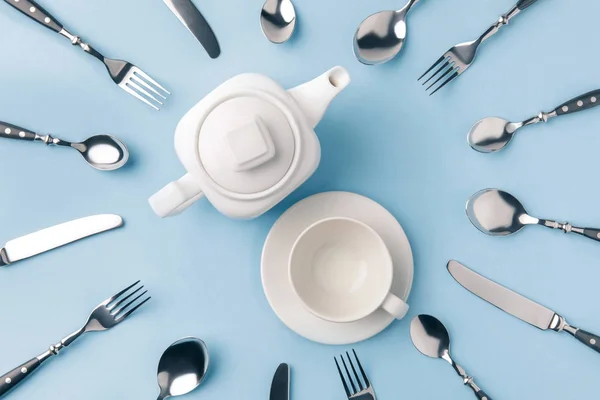 Vista superior de la tetera con taza en plato, rodeada de cubiertos aislados en azul - foto de stock