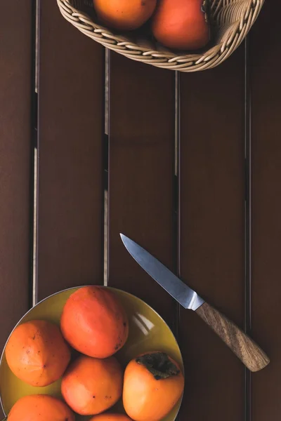 Top view of straw basket and plate with persimmons and knife on brown table — Stock Photo