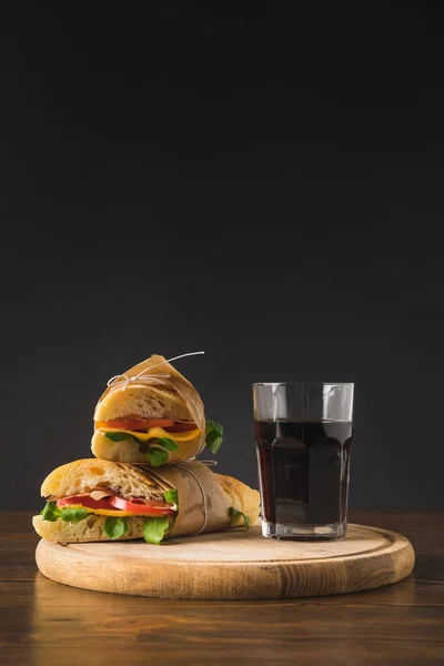Duas sanduíches uma sobre a outra e um copo de cola na tábua de corte — Fotografia de Stock