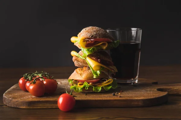 Sándwich grande con queso, tomates cherry y un vaso de cola en la tabla de cortar - foto de stock