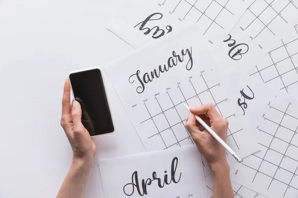 Visão parcial da mulher com smartphone na mão fazendo notas no calendário isolado no branco — Fotografia de Stock