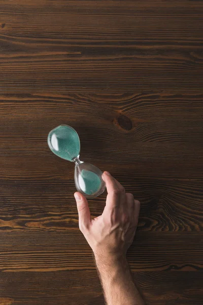 Partial view of man holding hourglass in hand on wooden surface — Stock Photo