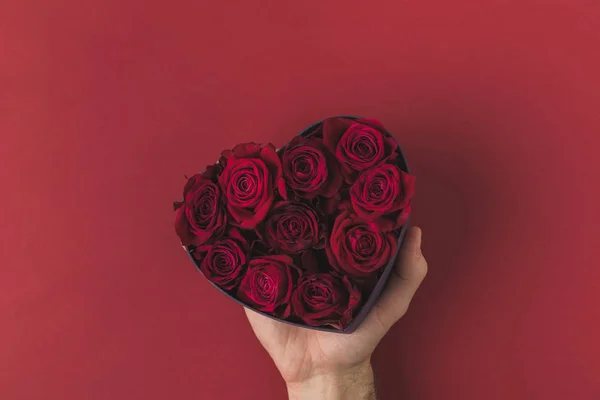 Cropped shot of man holding roses in heart shaped box on red tabletop, st valentines day concept — Stock Photo