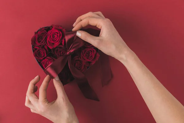 Partial view of woman tying ribbon on heart shaped box with roses on red tabletop, st valentines day concept — Stock Photo