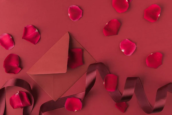 Плоска лежала з влаштованими пелюстками червоних троянд, конвертом і стрічкою ізольована на червоному, концепція дня Святого Валентина — стокове фото