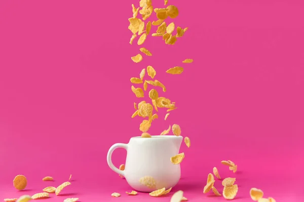 Крупный план хрустящих кукурузных хлопьев, падающих в молочный кувшин на розовый — стоковое фото