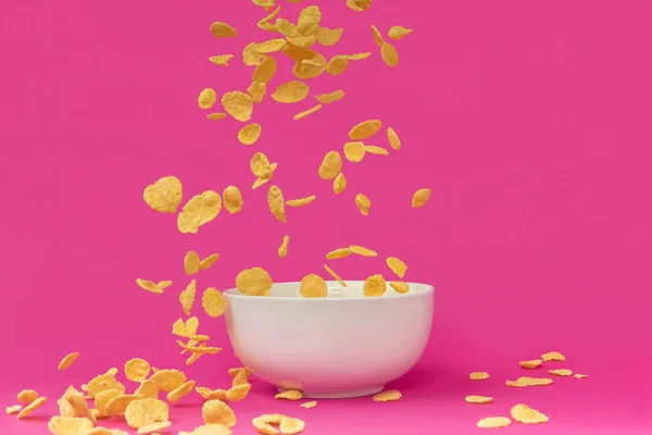 Крупным планом вид вкусных хрустящих кукурузных хлопьев, падающих в белую чашу, изолированную на розовый — стоковое фото