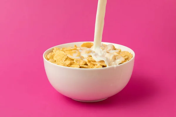 Nahaufnahme von frischer Milch in Schüssel mit Cornflakes auf rosa isoliert gießen — Stockfoto