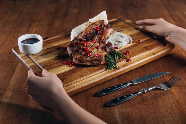 Cropped shot of woman holding grilled steak served with sauce and pomegranate seeds on wooden board — Stock Photo