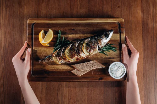 Tiro cortado de mulher segurando peixe grelhado com limão na tábua de madeira — Fotografia de Stock