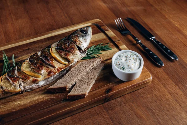 Close-up shot of grilled fish with lemon and sauce on wooden board — Stock Photo