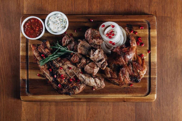 Varias carnes a la parrilla servidas sobre tabla de madera con salsas - foto de stock