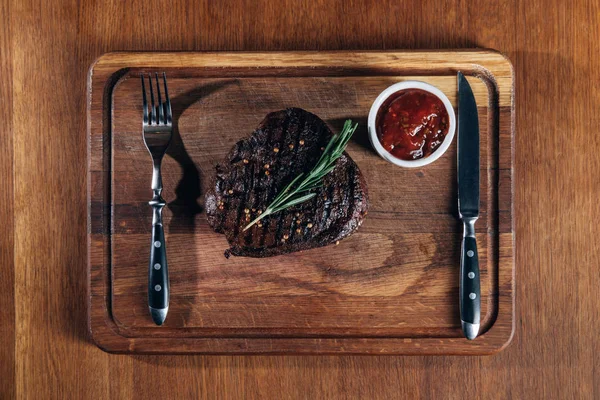 Vue du dessus du steak grillé avec couverts et sauce sur planche en bois — Photo de stock