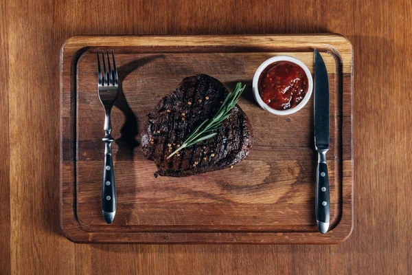 Vista superior de bistec a la parrilla con cubiertos y salsa sobre tabla de madera - foto de stock