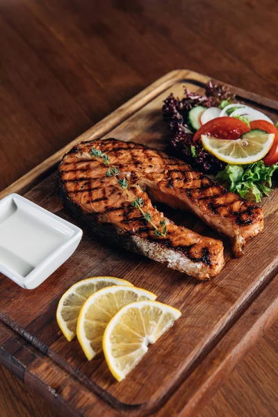 Close-up shot of grilled salmon steak served on wooden board with lemon slices and salad — Stock Photo