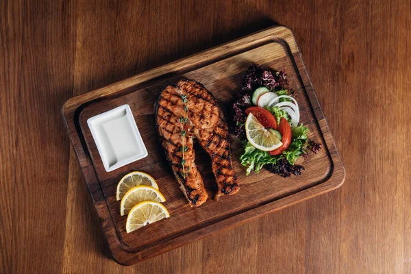 Vista superior de delicioso filete de salmón a la parrilla servido en tablero de madera con rodajas de limón y ensalada - foto de stock