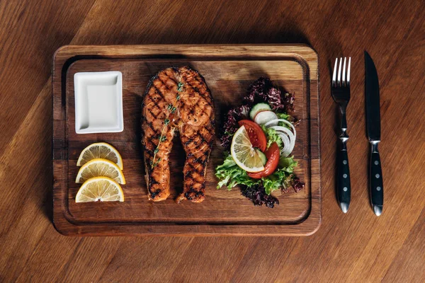 Vue du dessus du steak de saumon grillé servi sur une planche de bois avec tranches de citron et salade — Photo de stock