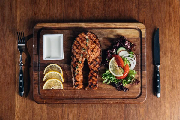 Вид зверху смачний смажений стейк з лососем, який подається на дерев'яній дошці з лимоном та салатом — стокове фото