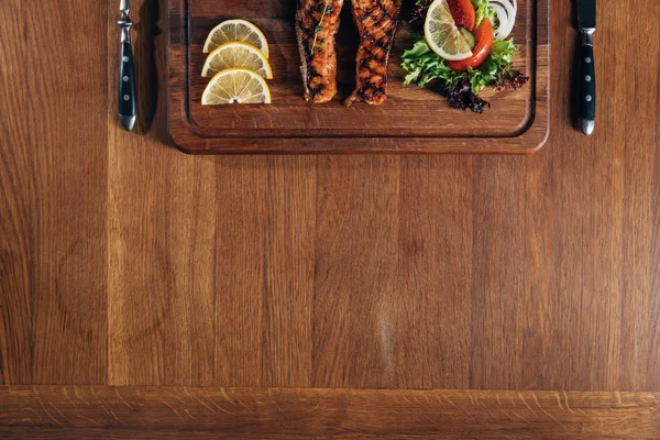 Вид на вкусный стейк из лосося на гриле, подаваемый на деревянной доске с лимоном и салатом — стоковое фото
