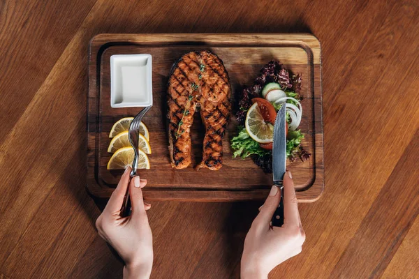 Cropped shot of woman eating grilled salmon steak served on wooden board with lemon and lettuce — Stock Photo