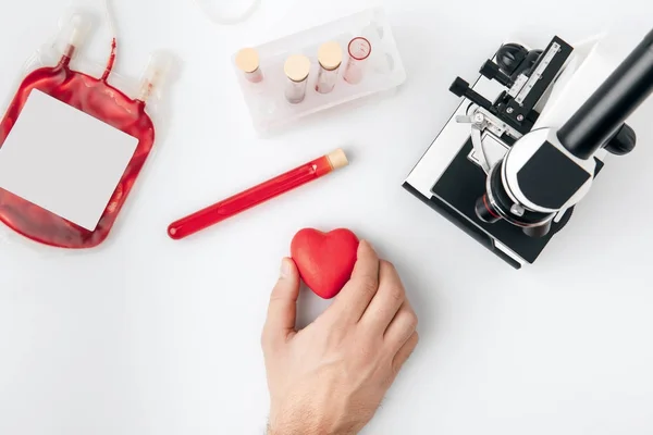 Top view of hand holding red heart against vials with blood and microscope isolated on white background — Stock Photo