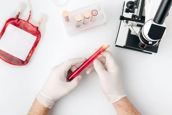 Vista superior das mãos em luvas segurando sangue para transfusão em frasco para injetáveis e microscópio isolado em fundo branco — Fotografia de Stock