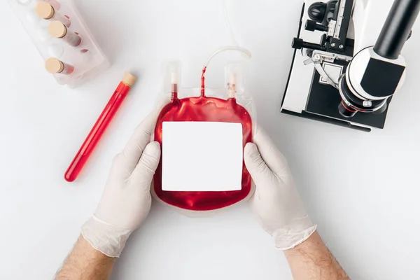 Vista dall'alto delle mani in guanti che tengono il sangue per trasfusione vicino ai flaconcini e microscopio isolato su fondo bianco — Foto stock