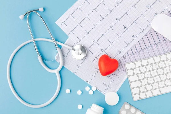 Estetoscopio, papel con cardiograma, pastillas dispersas, corazón rojo y teclado aislados sobre fondo azul - foto de stock