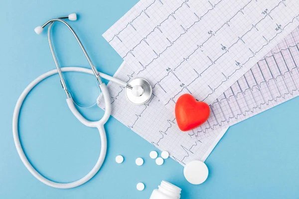 Stethoskop, Papier mit Kardiogramm, verstreute Pillen und rotes Herz isoliert auf blauem Hintergrund — Stockfoto