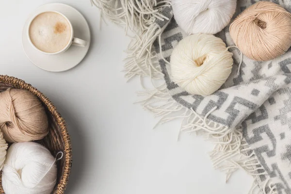 Vista dall'alto di palline di filato in cesto di vimini, tazza di caffè e coperta su sfondo bianco — Foto stock