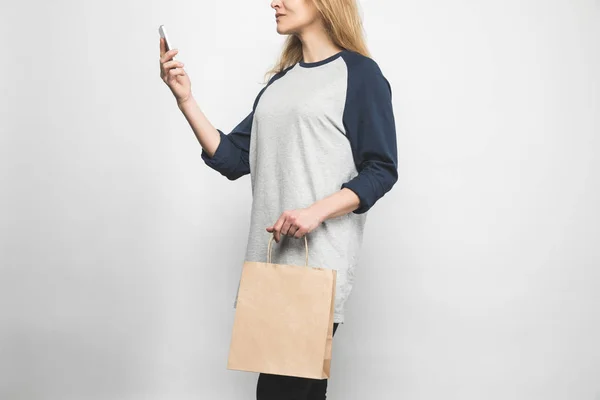 Cropped shot of woman in stylish long sleeve on white with shopping bag and smartphone — Stock Photo