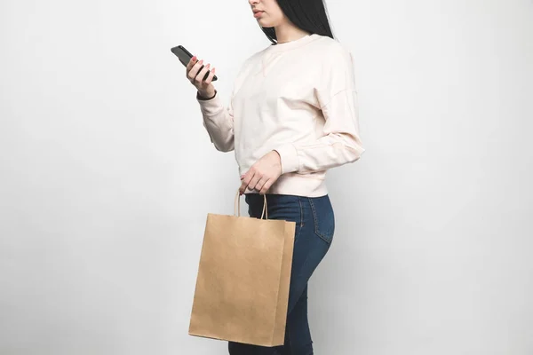 Cropped shot of young woman in blank sweatshirt on white with shopping bag and smartphone — Stock Photo