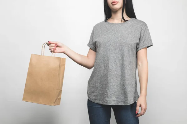 Cropped shot of young woman in blank grey t-shirt on white with shopping bag — Stock Photo