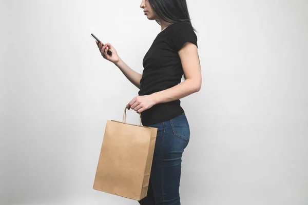 Cropped shot of young woman in blank black t-shirt isolated on white with shopping bag and smartphone — Stock Photo