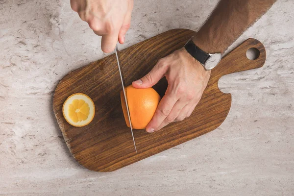 Imagen recortada de hombre cortando naranja en tablero de madera - foto de stock