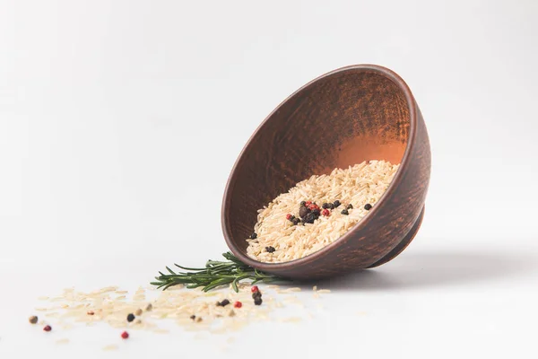 Raw rice and peppercorns spilling out bowl on white surface — Stock Photo