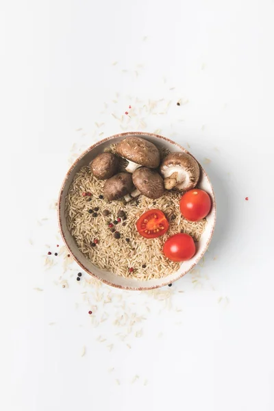 Top view of raw rice in bowl with mushrooms and tomatoes on white surface with spilled rice and spices around — Stock Photo