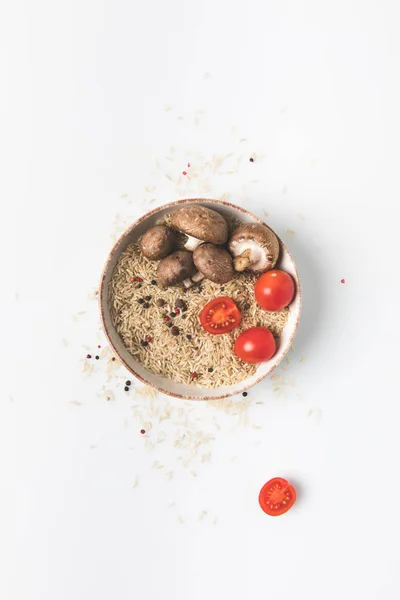 Vista dall'alto del riso crudo in ciotola con funghi e pomodori su tavolo bianco con riso versato e spezie intorno — Foto stock