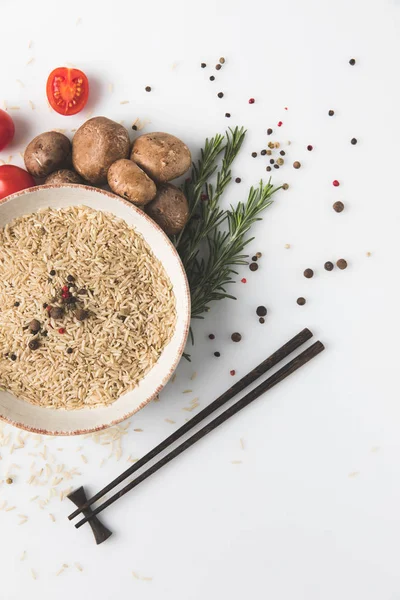 Vista superior de arroz cru em tigela com cogumelos e tomates na superfície branca com pauzinhos — Fotografia de Stock