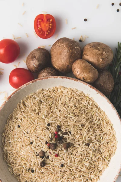 Top view of raw rice and spices in bowl with mushrooms and tomatoes on white surface — Stock Photo