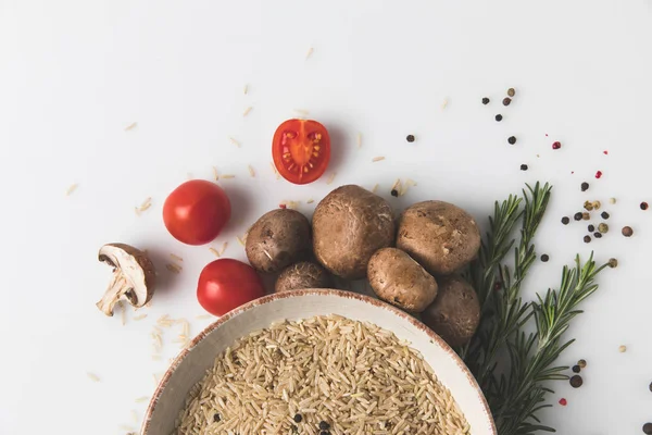 Vista superior del arroz crudo en tazón con champiñones y tomates en la superficie blanca - foto de stock