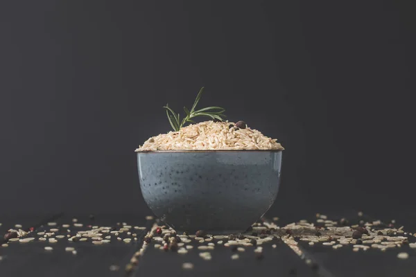 Schüssel mit rohem Reis mit Rosmarin und Gewürzen auf schwarzer Oberfläche — Stockfoto