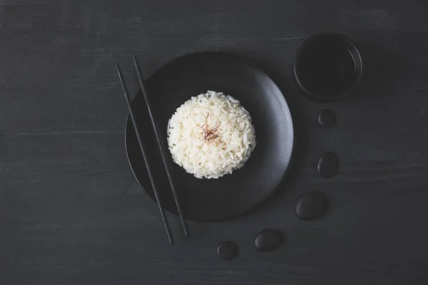 Vue du dessus de riz savoureux sur assiette avec baguettes sur table noire — Photo de stock