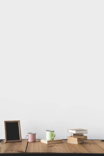 Empty blackboard in frame with cups and books on wooden table on white — Stock Photo