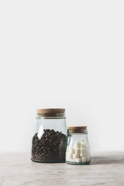 Granos de café y azúcar refinada en botellas de vidrio sobre mesa de mármol sobre blanco - foto de stock